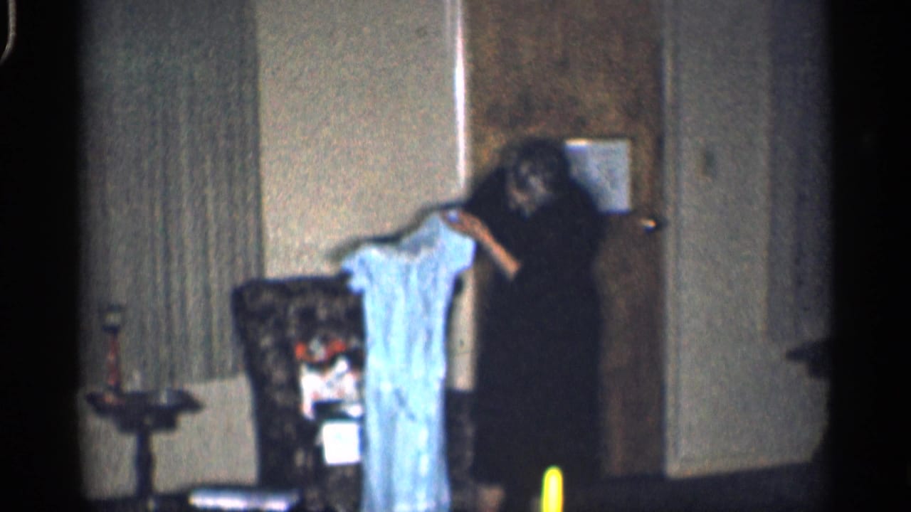 Woman continues holding a blue dress beside a door in a dim living room.
