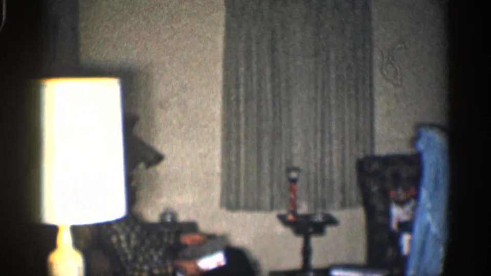 A bright lamp dominates the foreground while a seated man in plaid is partly visible near the chair and blue dress.