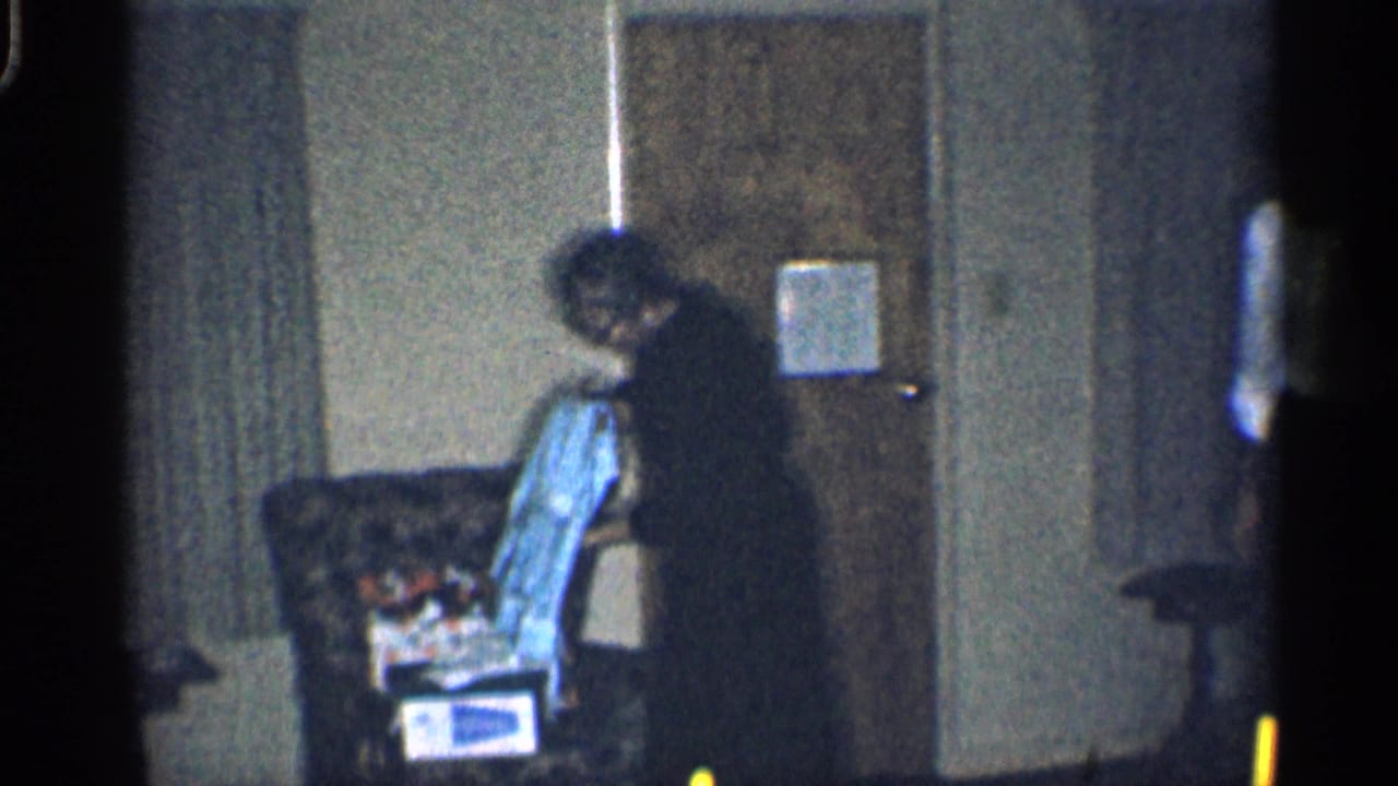 Woman in a dark dress bends near an armchair with clothing in a living room.