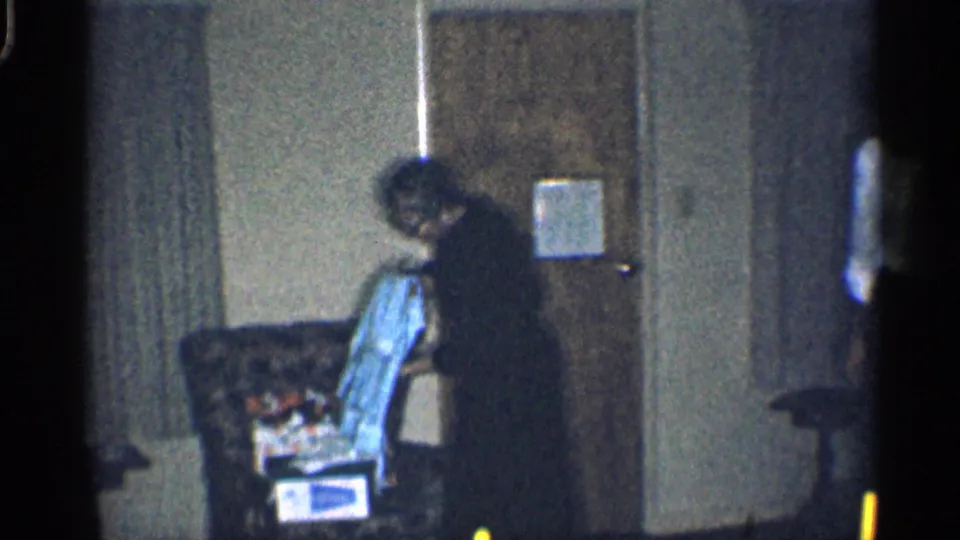 1959 California home-movie frame showing a woman in a dark dress arranging clothing on a patterned armchair in a living room.