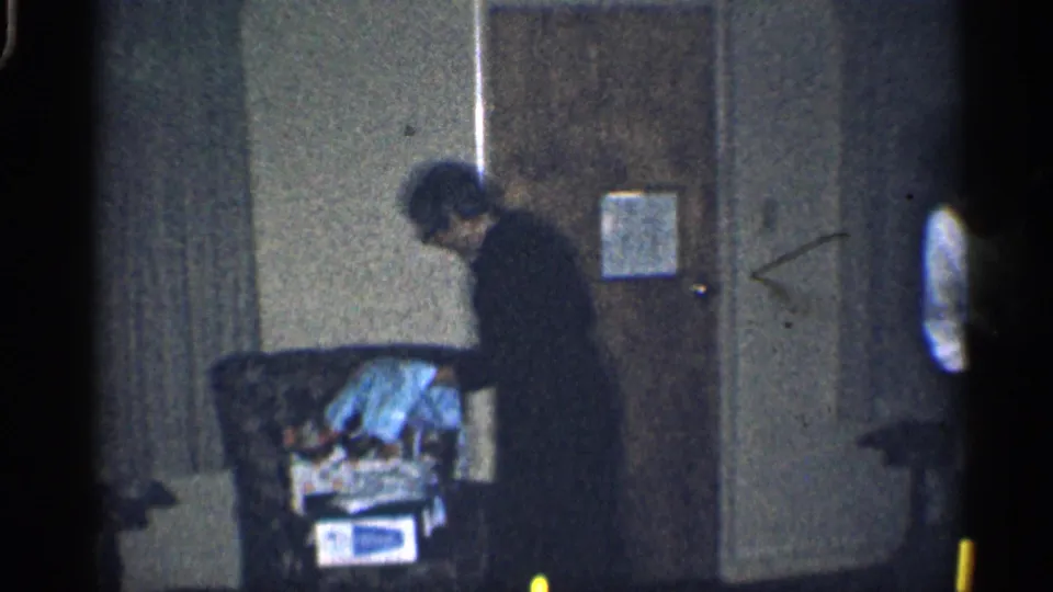 1959 California home-movie frame showing a woman beside a chair with clothing, with another person partly visible at the room edge.