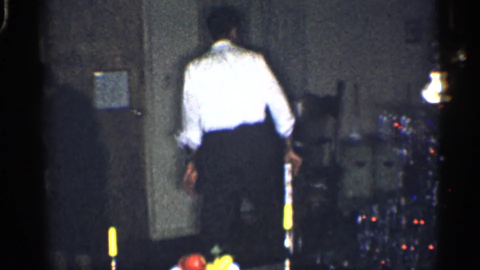 1959 California home-movie frame showing a person in a white shirt walking through a dim living room with a lamp and decorated tree.