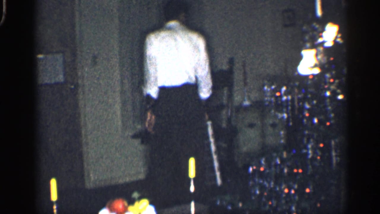 In 1959 California home-movie footage, a man in a white shirt stands near a lit Christmas tree in a dim private interior.