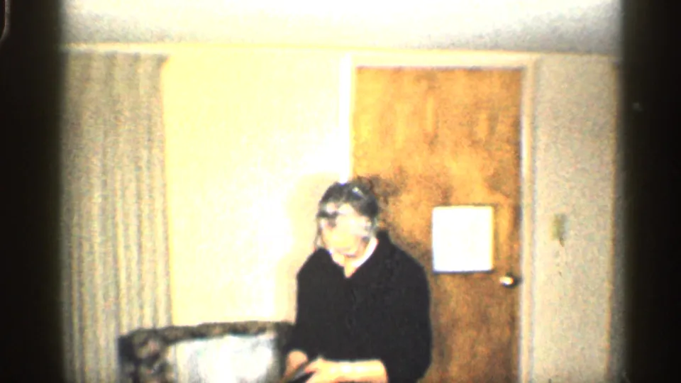 An older woman sits reading near curtains and a wooden door in a private California home interior from the 1959 footage.