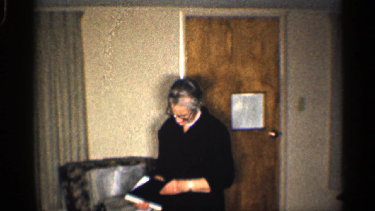 Woman with glasses reading a book or document in a modest living room.