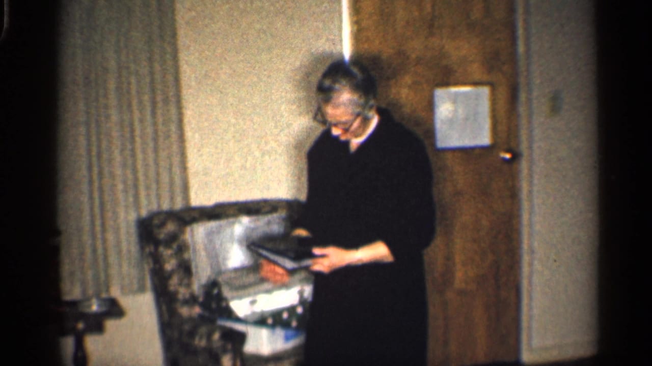 Woman standing indoors with an open book or document near an armchair.