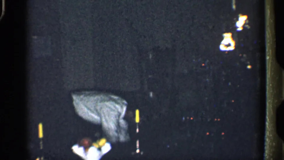A low-light 8mm nighttime scene shows an indistinct figure bending near small bright points of light, with details obscured by darkness and grain.