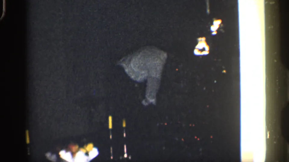 Low-light 8mm frame from 1959 California showing an adult man leaning downward in a shadowy interior.