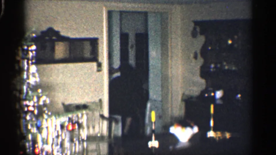 Low-lit 1959 California holiday interior showing a tinsel Christmas tree, doorway, wall shelf, candles, and surrounding furniture.