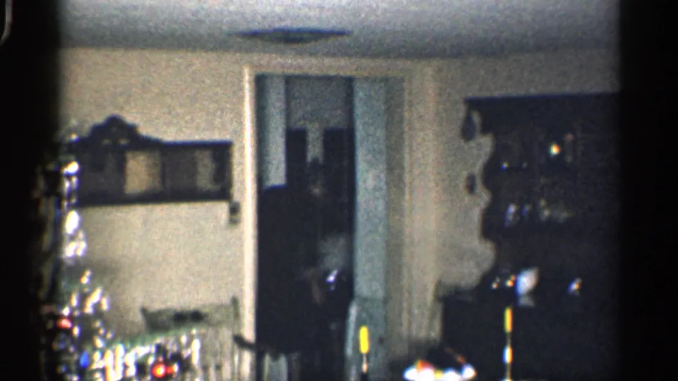 1959 California home-movie frame showing a dim living room before the man enters, with a doorway, cabinet, candles, and holiday decor.