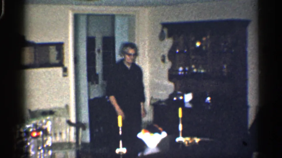 1959 California home-movie frame showing an older man in dark clothing and glasses entering a living room with holiday decor.