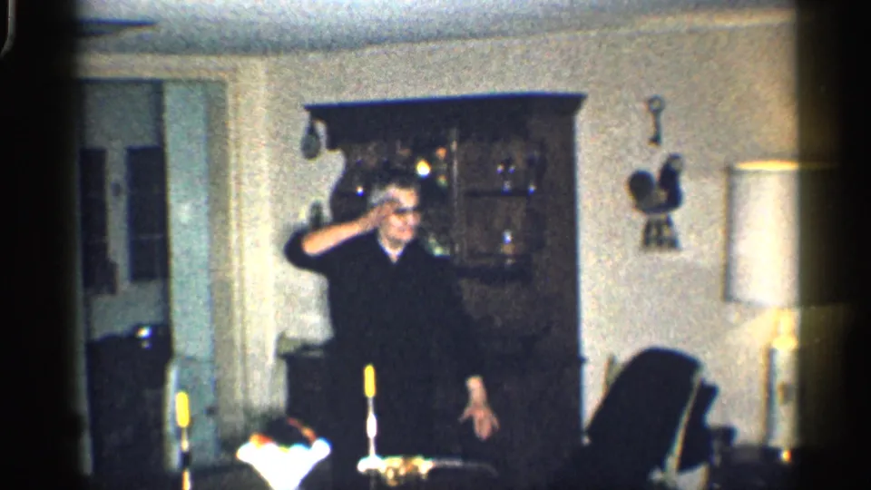 1959 California home-movie frame of an older man in glasses making a playful hand gesture in a dim living room.
