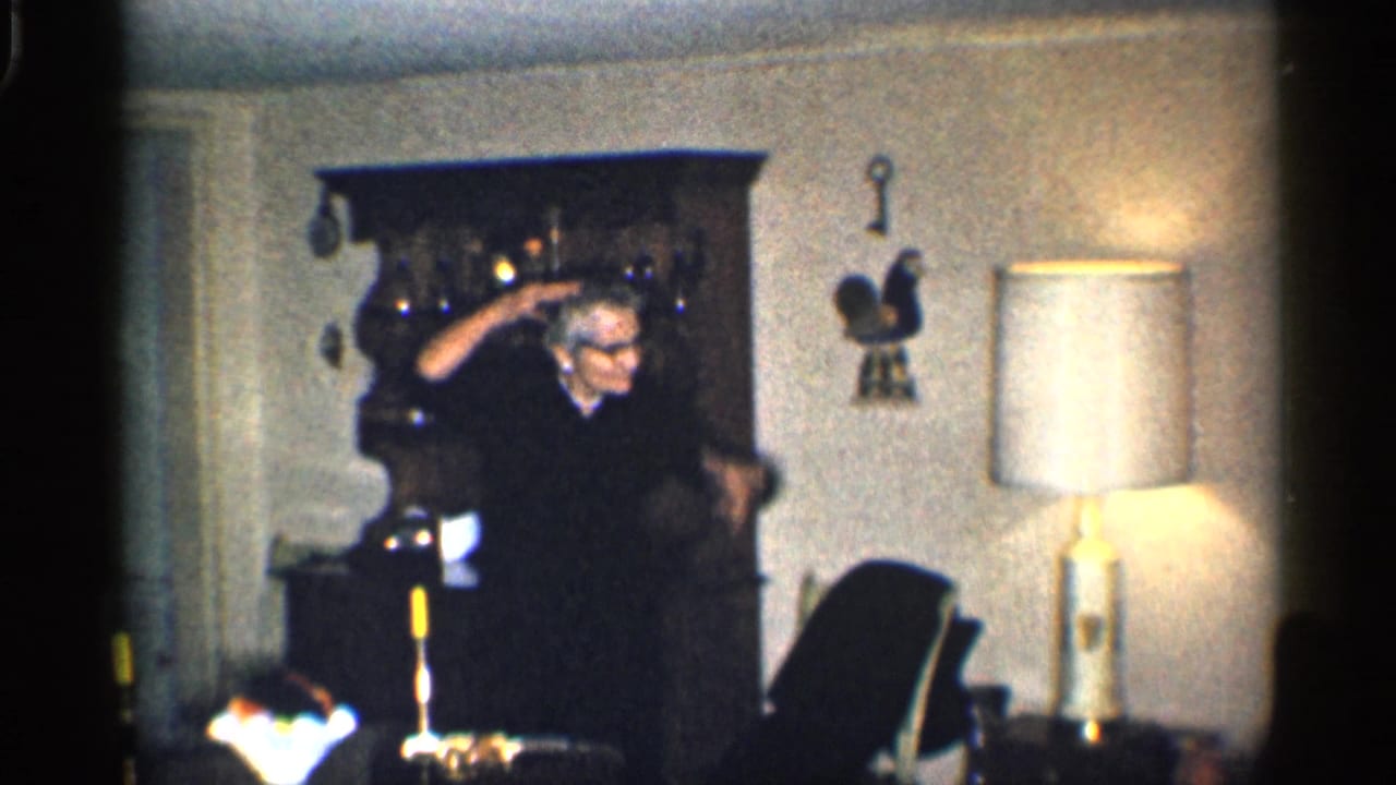 Adult in a dark sweater gestures beside a wooden hutch in a living room.