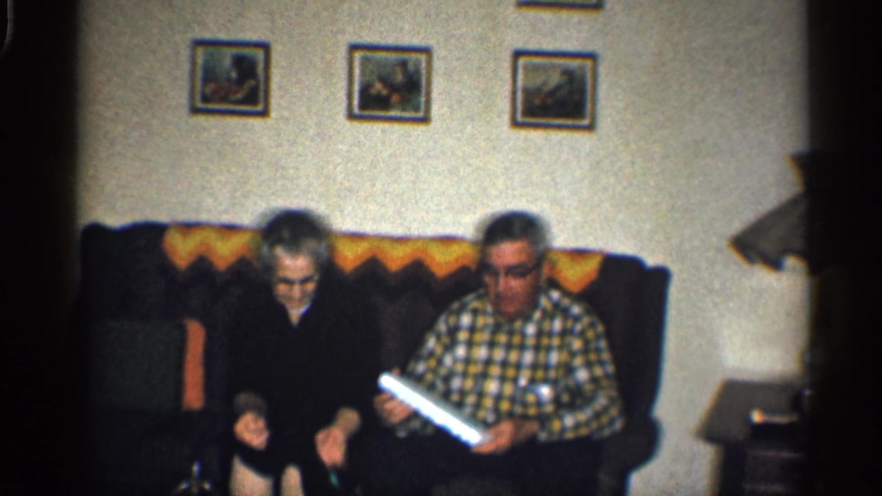 Two adults sit on a dark couch while one holds a glowing rectangular object.