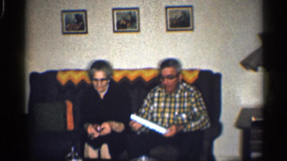1959 California home-movie frame showing two adults seated on a couch, with a patterned throw, framed pictures, and a side table visible.