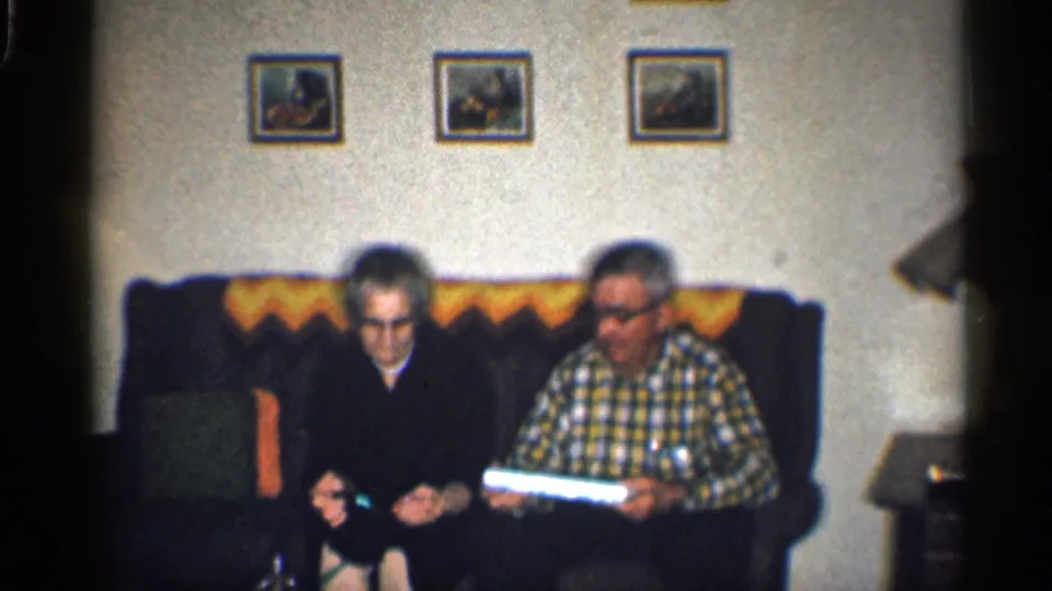 1959 California living-room frame with two adults seated on a couch, framed pictures above, and a bright rectangular object in view.