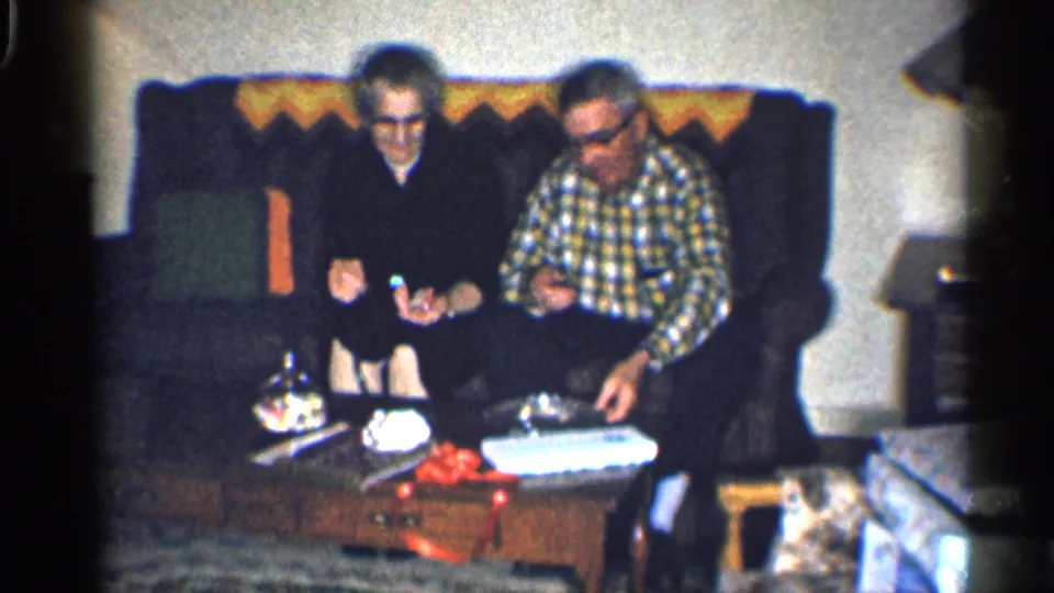 The couple sits close together as gifts, a white box, and small objects rest on the coffee table in front of them.