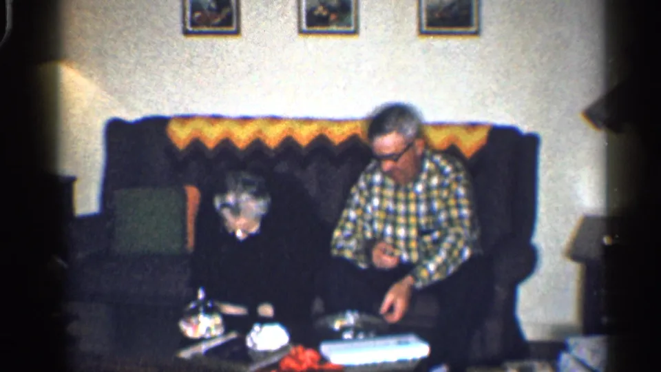 At the end of the clip, the woman bends toward the coffee table while the man studies a small object on the sofa.