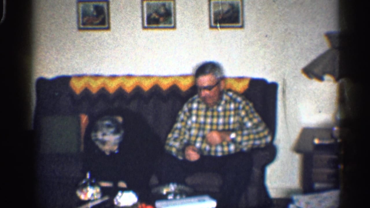 Older man seated on a dark couch in a living room with framed pictures above.