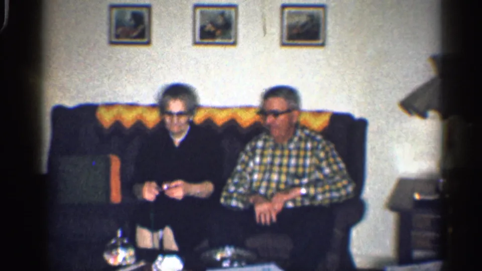 1959 California home-movie frame showing an older woman and older man seated together on a couch in a private living room.