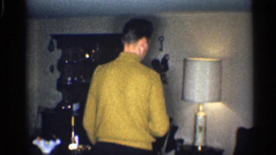 1959 California home-movie frame of a man in a yellow sweater viewed from behind in a private interior with a table lamp.