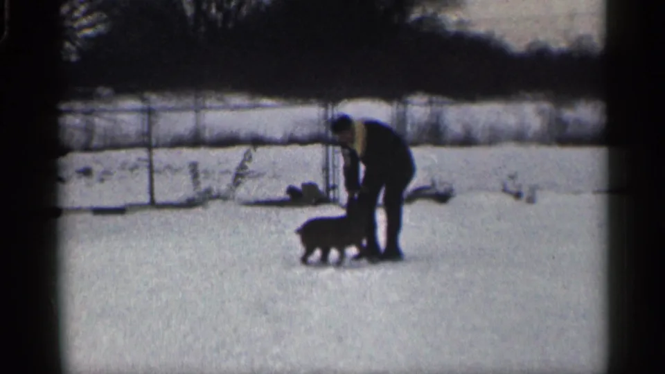 A 1959 home-movie frame shows the man crouching or bending near two dogs in a snow-covered fenced yard.