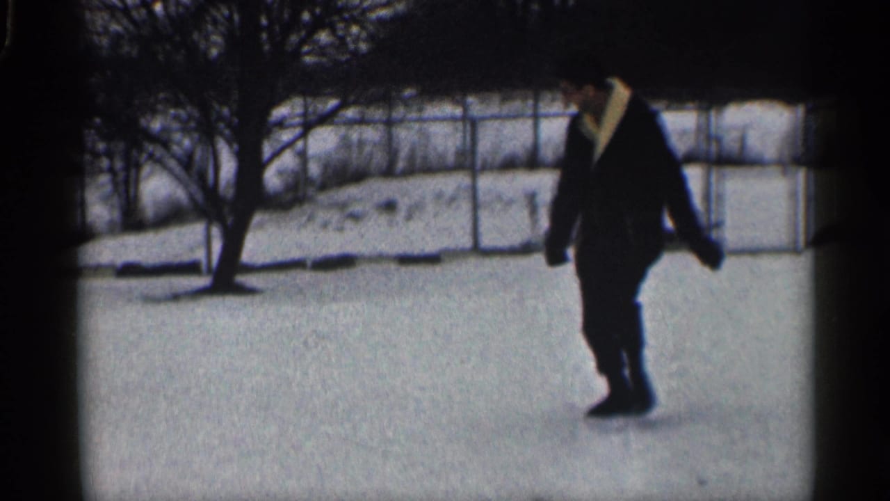 Man in a dark coat walking across a snowy yard near bare trees and a fence.