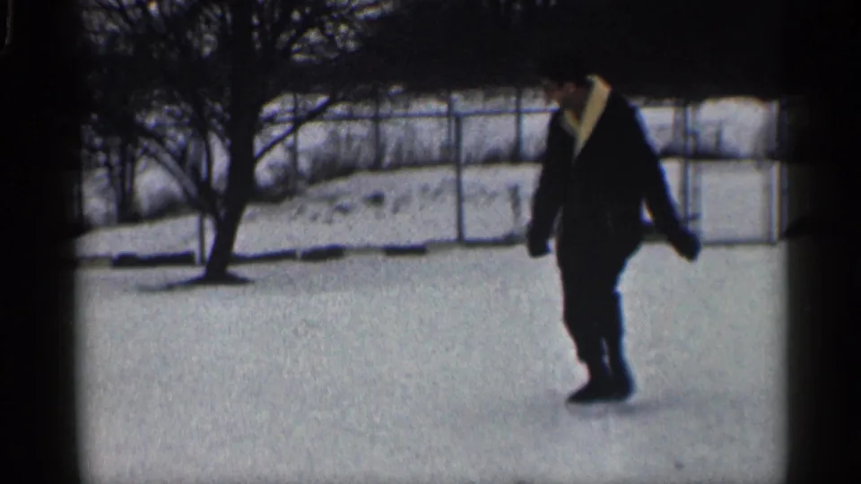 In a 1959 California home-movie frame, a man in a dark coat walks across snow with bare trees and a chain-link fence behind him.