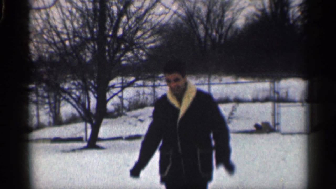 Man in a dark coat approaches the camera across a snowy yard.