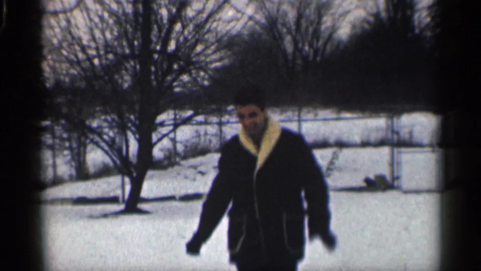 The man walks toward the camera through a snowy California yard, with bare trees and fencing visible in the background.