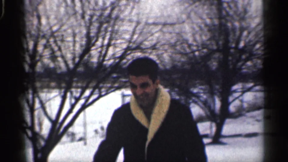 A closer home-movie view shows the man in a dark coat and light collar near bare winter branches in the snowy yard.