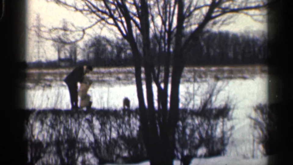 1959 California 8mm frame showing a distant man and dark dogs near bare trees, snow, and a frozen-looking body of water.