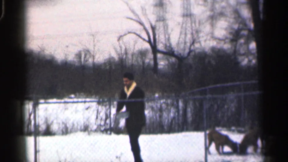 A man in a dark coat and hat walks beside a chain-link fence in a snowy California yard, with bare trees behind him.