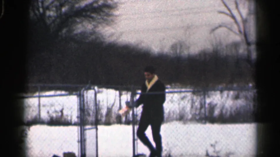 The bundled man pauses in the snowy fenced yard, holding a light object as bare trees and brush fill the background.