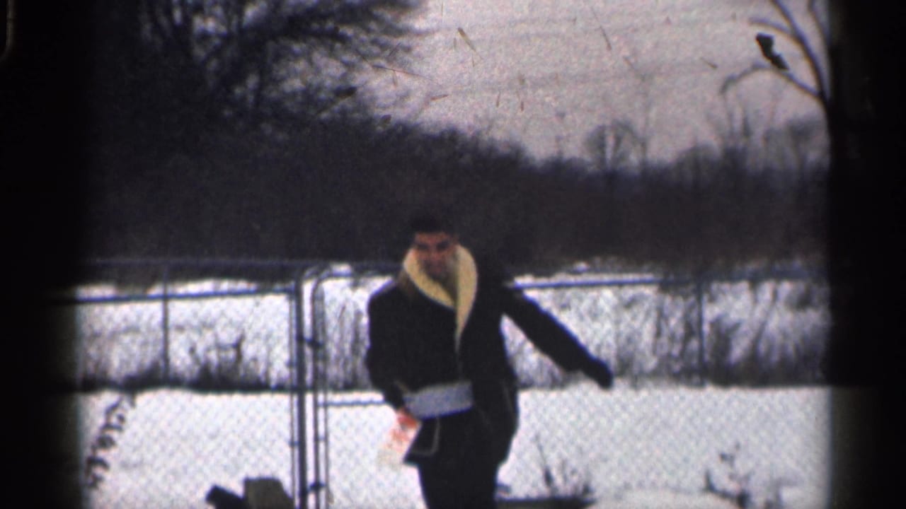In 1959 California home-movie footage, a man in a dark coat with a light collar walks across a snowy yard near a fence.