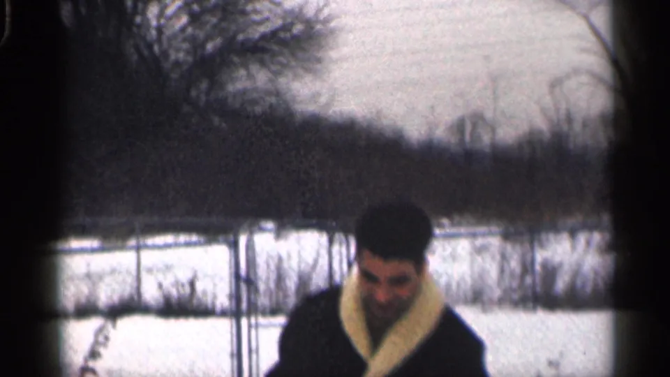 A man in a dark coat lowers his gaze while walking through a snowy yard, with bare trees and fencing behind him.