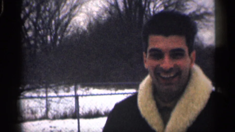 Close home-movie view of the smiling man in a dark coat with a light collar, framed against snow and winter trees.
