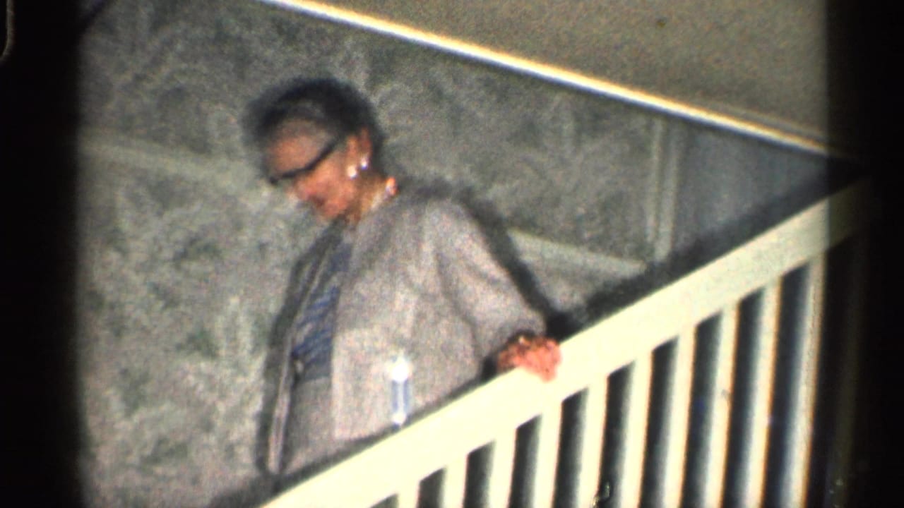 Elderly woman in glasses holds a white stair railing inside a home.