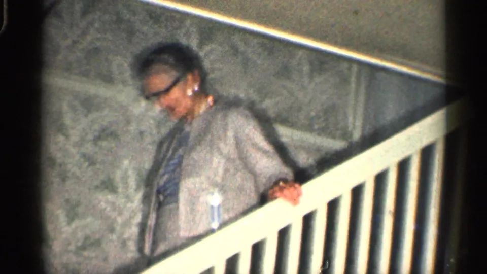 In 1959 California home-movie footage, an elderly woman in glasses descends an indoor staircase beside patterned wallpaper.