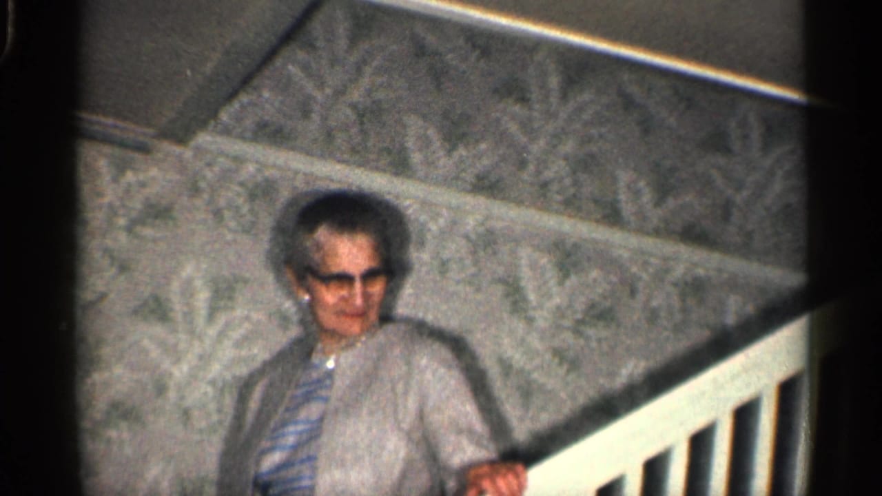 Woman in glasses stands by the stair railing against patterned wallpaper.