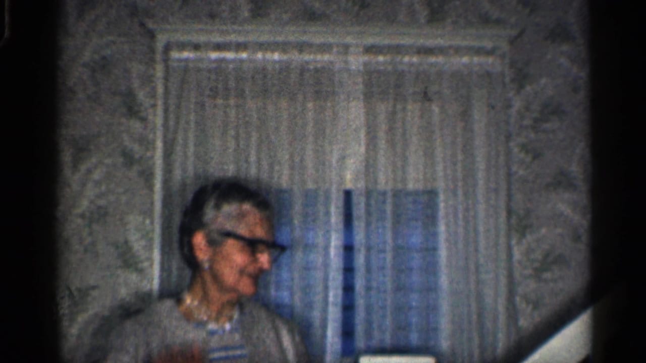 Older woman in dark-rimmed glasses inside a California home, with sheer curtains and patterned wallpaper behind her.