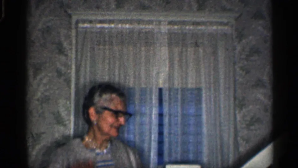 Older woman in dark-rimmed glasses inside a California home, with sheer curtains and patterned wallpaper behind her.