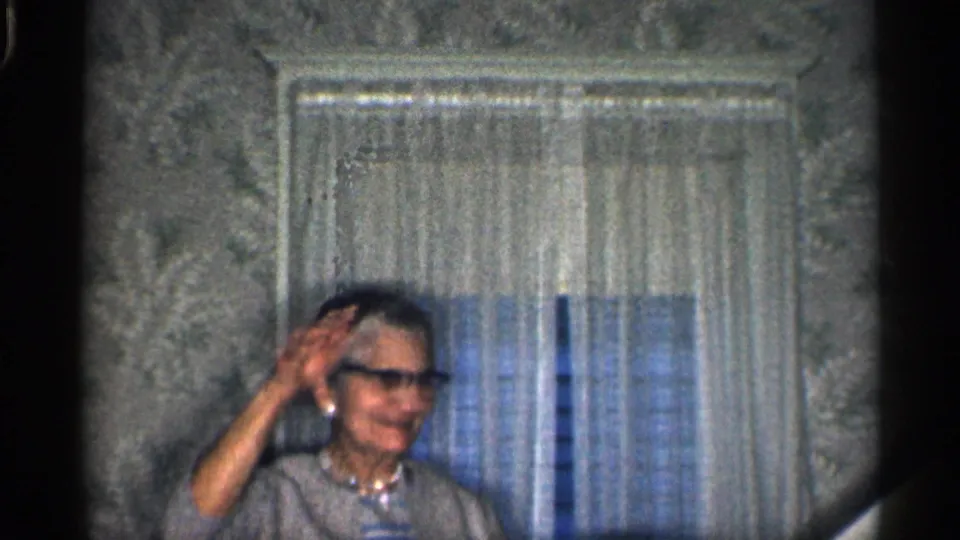 Older woman in glasses waves toward the camera in a 1959 California home interior with curtains and wallpaper.