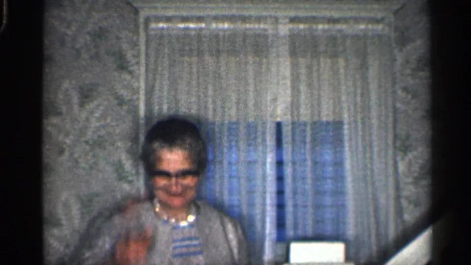 Older woman in glasses looks down during a small wave inside a California home, framed by curtains and patterned wallpaper.