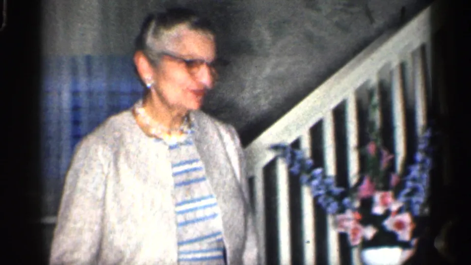 Older woman in a light cardigan stands near a staircase and flowers in a 1959 California home interior.