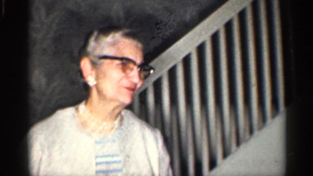 In 1959 California home-movie footage, an elderly woman in dark-rimmed glasses, pearls, cardigan, and striped dress smiles near a staircase.