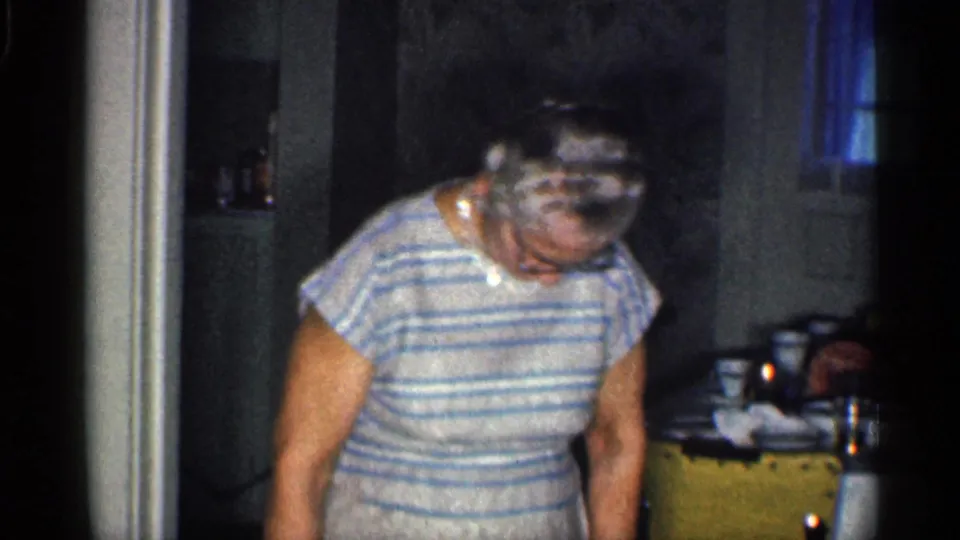 The woman appears in a striped dress and pearls inside a California home, looking down near a doorway with household items in the background.