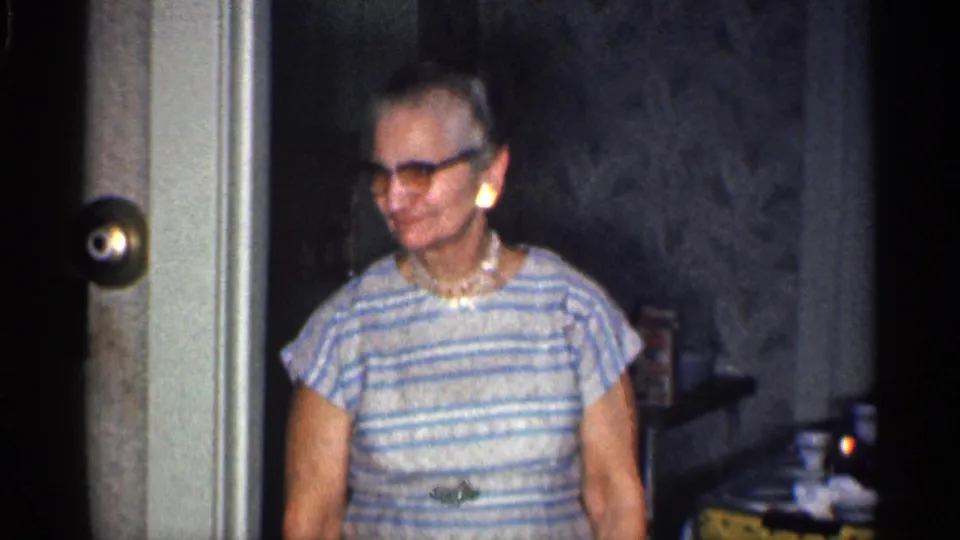 A 1959 California home-movie frame shows the elderly woman smiling in dark-rimmed glasses and a striped dress near a doorway and tableware.