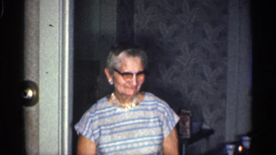The elderly woman smiles in a dim California home interior, wearing dark-rimmed glasses, pearls, and a blue-and-white striped dress.