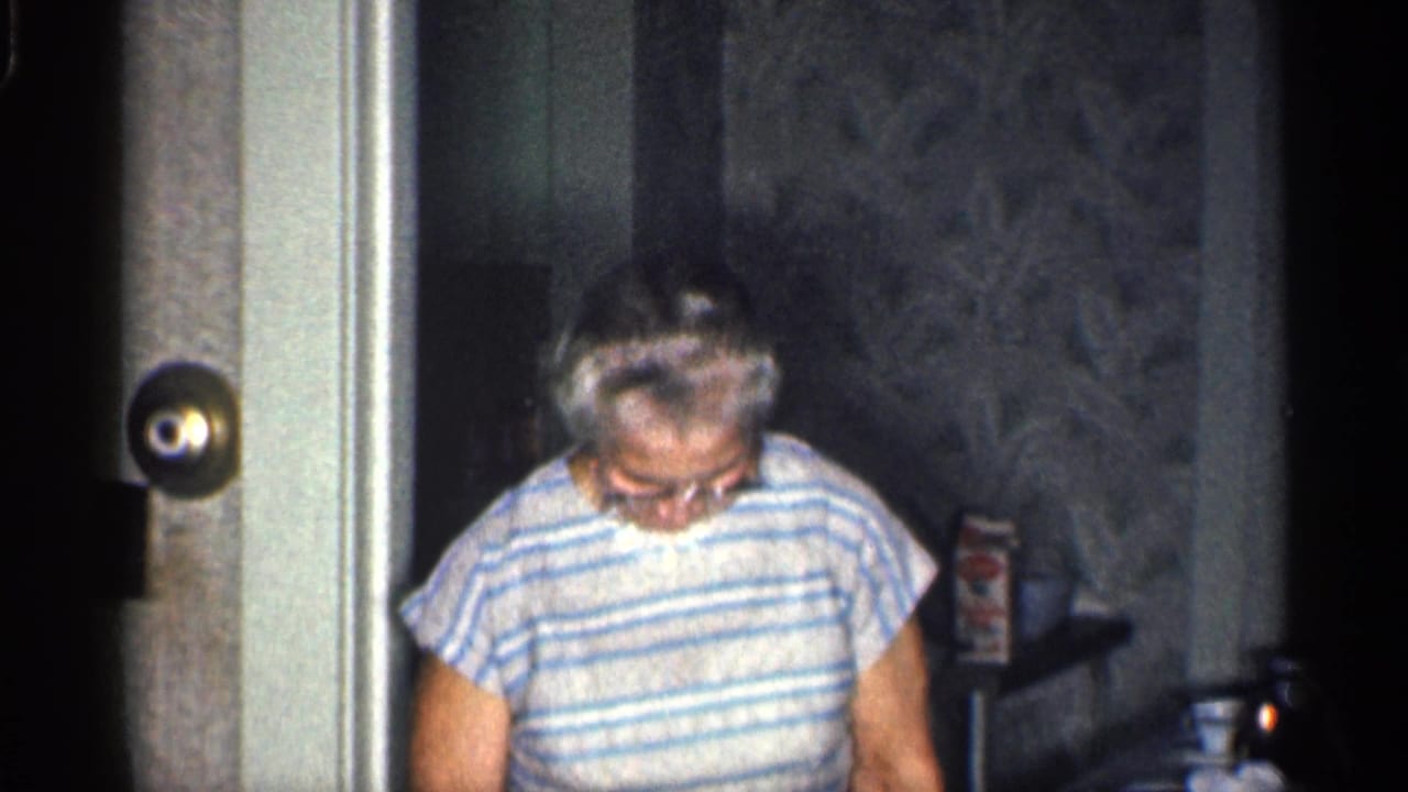 In a 1959 California home interior, an older woman in glasses and a striped top looks down while standing near a doorway.